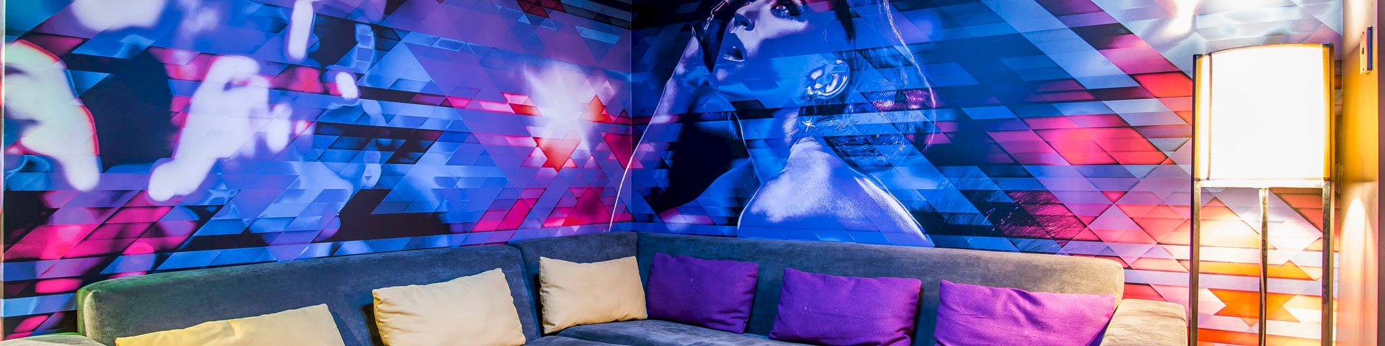 A colorful room with a large L-shaped sofa, vibrant wall art, ottomans, and a floor lamp under blue lighting.