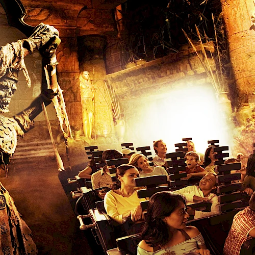 An image of people on a theme park ride, featuring a menacing skeleton wielding an axe in an ancient, temple-like setting.