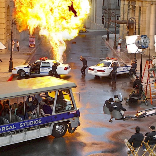 A movie set at Universal Studios with a controlled explosion, police cars, film crew working, and a tour bus with spectators watching the action.