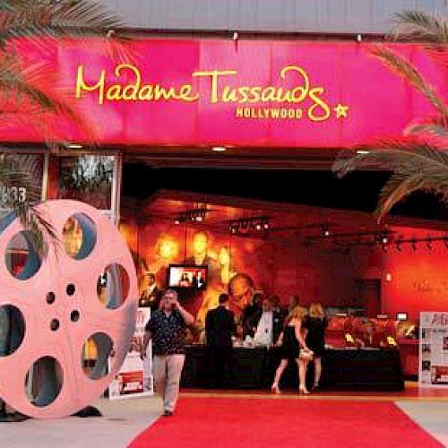 This image shows the entrance to Madame Tussauds Hollywood with a red carpet, palm trees, and large film reel props decorating the pathway.