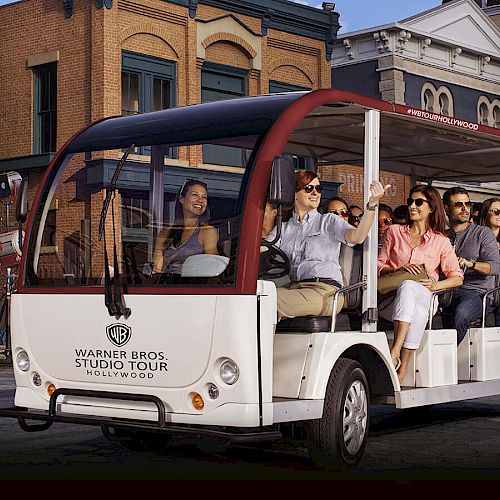 A group of people are riding in an open-air tram, taking a Warner Bros. Studio Tour in Hollywood, with buildings in the background ending the sentence.