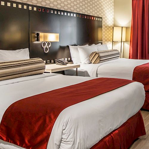 The image shows a hotel room with two double beds, red accents, decorative pillows, a lamp, and a window with curtains in the background.