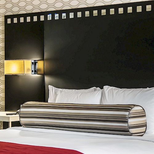 A modern hotel room features a large bed with white pillows and a striped cylindrical cushion, a bedside table, and a wall-mounted lamp ending the sentence.