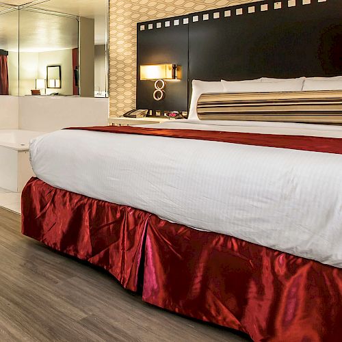 A hotel room features a luxurious bed with a red skirt and striped bolster pillow, a large hot tub, and a mirrored wall behind the tub.