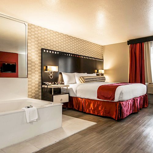 The image shows a hotel room with a bed, a jacuzzi, nightstands with lamps, and a window with curtains in a well-lit space.