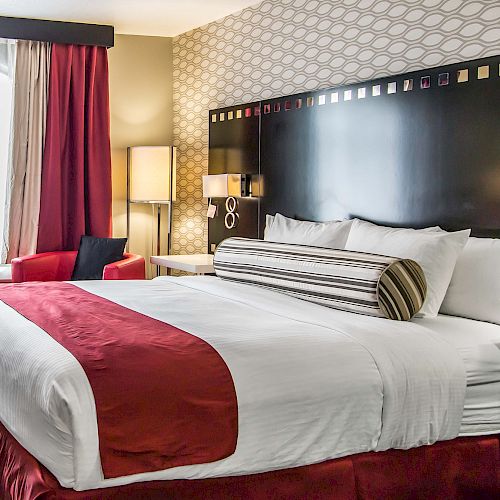 The image shows a hotel room with a neatly made bed, red and white linens, a headboard, a lamp, a window with curtains, and a small sitting area.