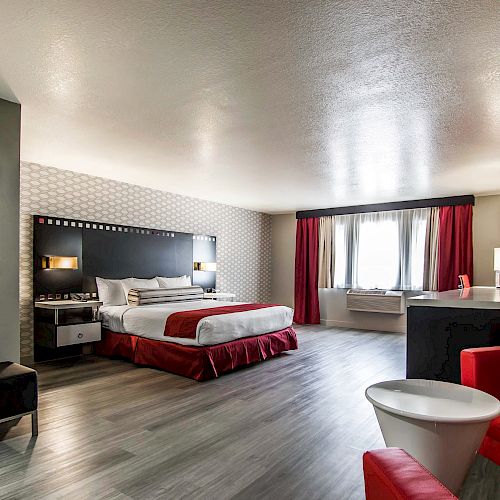 A modern hotel room with a king-sized bed, red accents, sitting area, and large windows allowing natural light to fill the space.