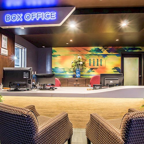 This image shows a modern box office with two chairs in the foreground, computers on the desk, and colorful wall art in the background.