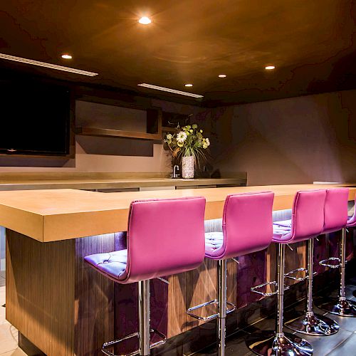 A modern bar with a wooden countertop, purple stools, a wall-mounted TV, and a vase with flowers on the counter under warm lighting.