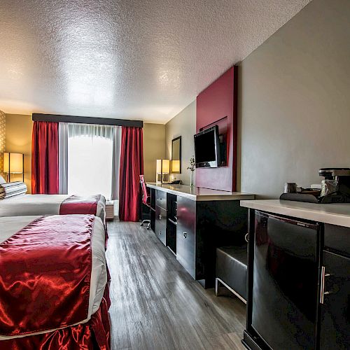 A hotel room with two beds, a TV, desk, coffee maker, and a window with red curtains at the far end of the room is shown.