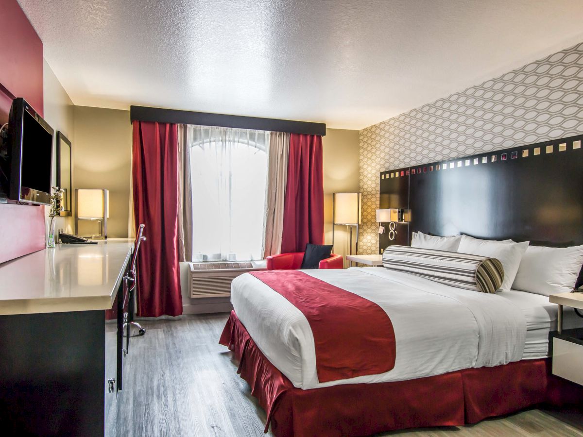 The image shows a modern hotel room with a large bed, red and white bedding, a TV, a desk, lamps, and a window with red and white curtains.