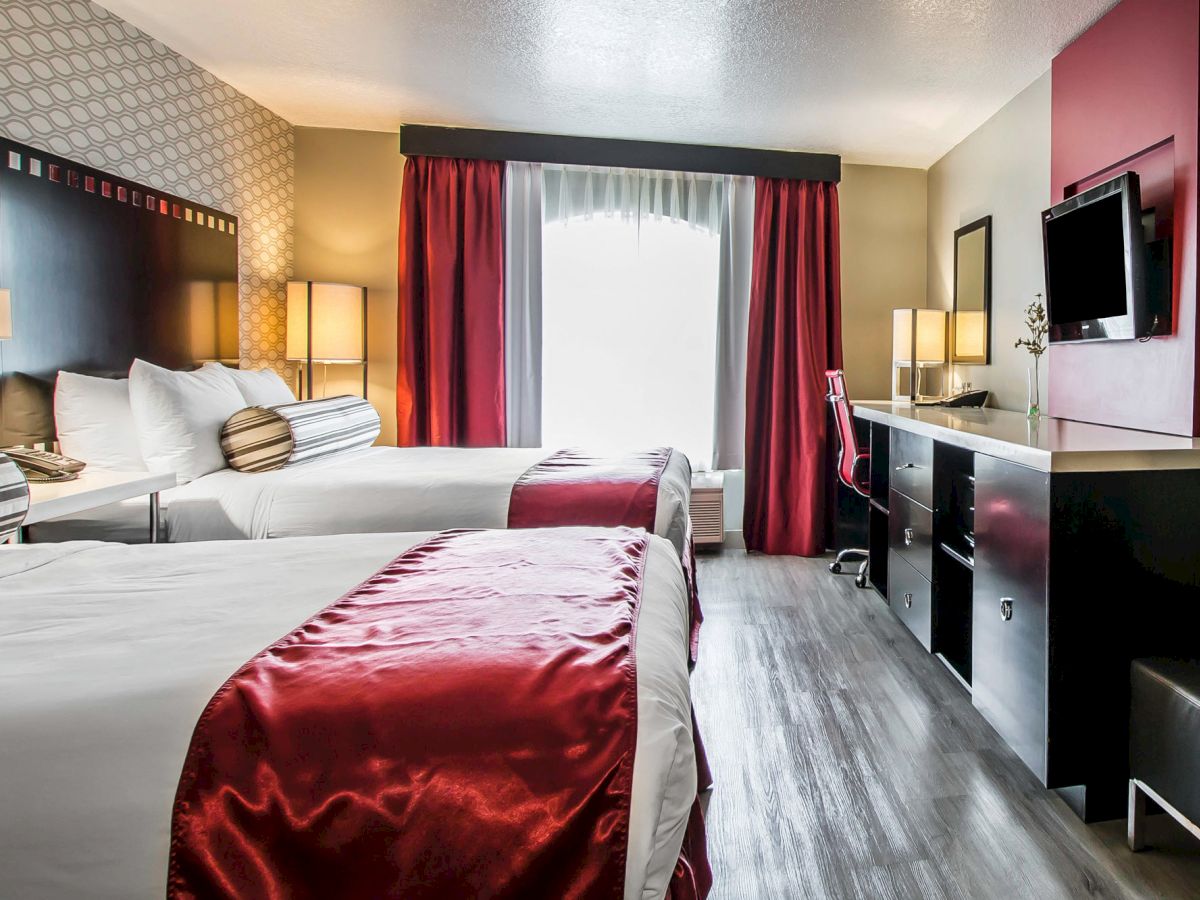 This image shows a modern hotel room with two double beds, each with white linens and red runners. There's also a desk, a TV, and red curtains.