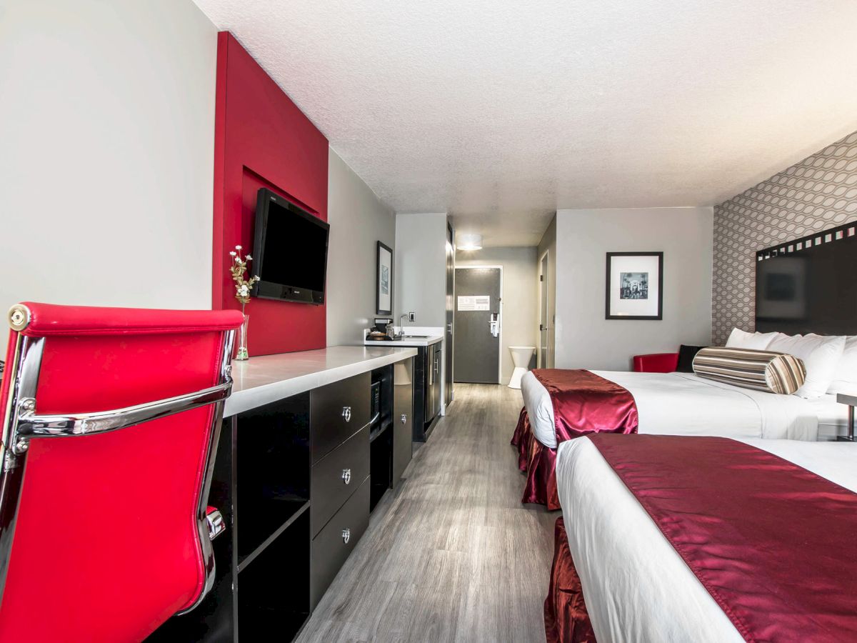 A modern hotel room with two double beds, a red-accented chair, flat-screen TV, wall art, and a minimalist desk area.