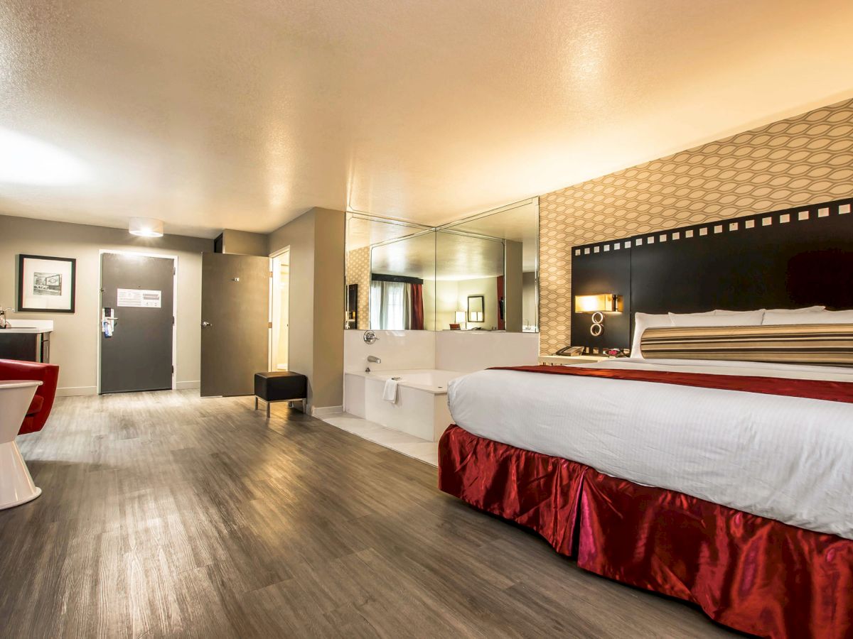 A spacious hotel room with a large bed, red accents, a desk, and a mirror, featuring modern decor and ample lighting.