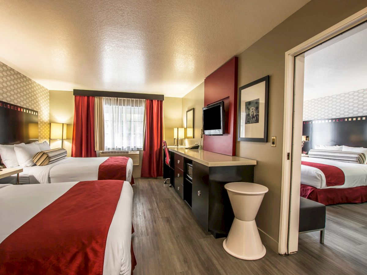 The image shows a hotel room with two beds featuring red accents, a second behind a partially open divider, a TV, a desk, and modern decor.