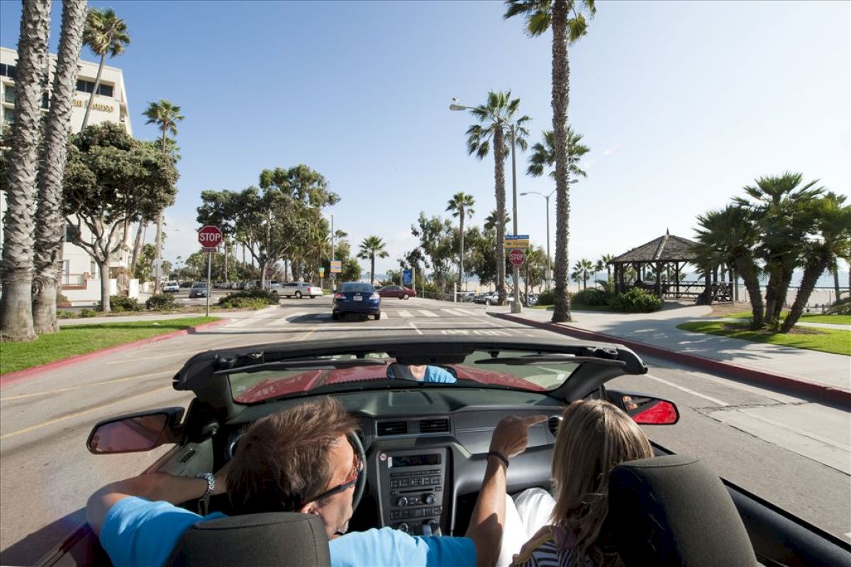 A man and woman driving in a red convertible on a sunny, palm-lined street by the beach, with a gazebo visible in the background.