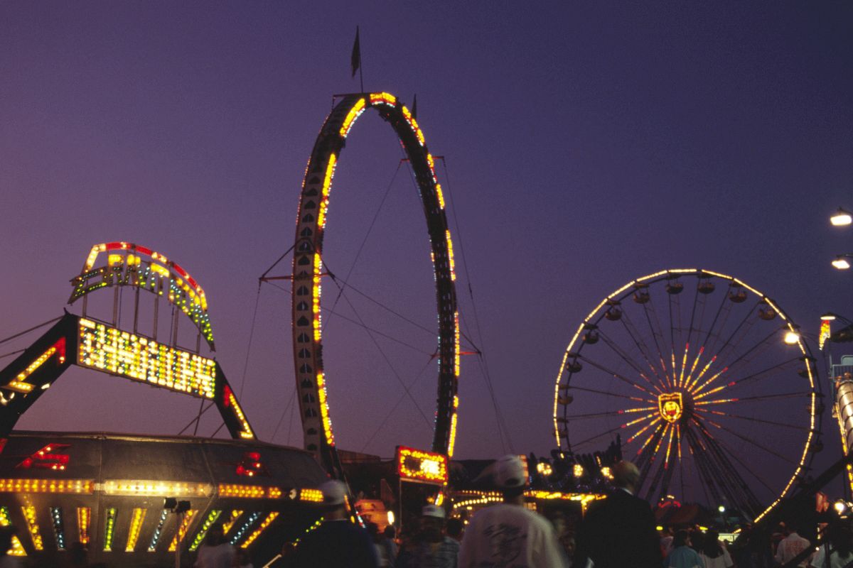 The image shows a night scene at a fair with illuminated rides, including a Ferris wheel, a circular ride, and a roller coaster, with people below.