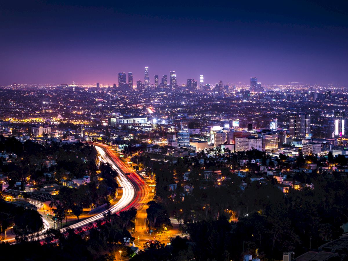 A nighttime cityscape featuring a well-lit urban area with a prominent highway and tall buildings in the background, under a dark sky.