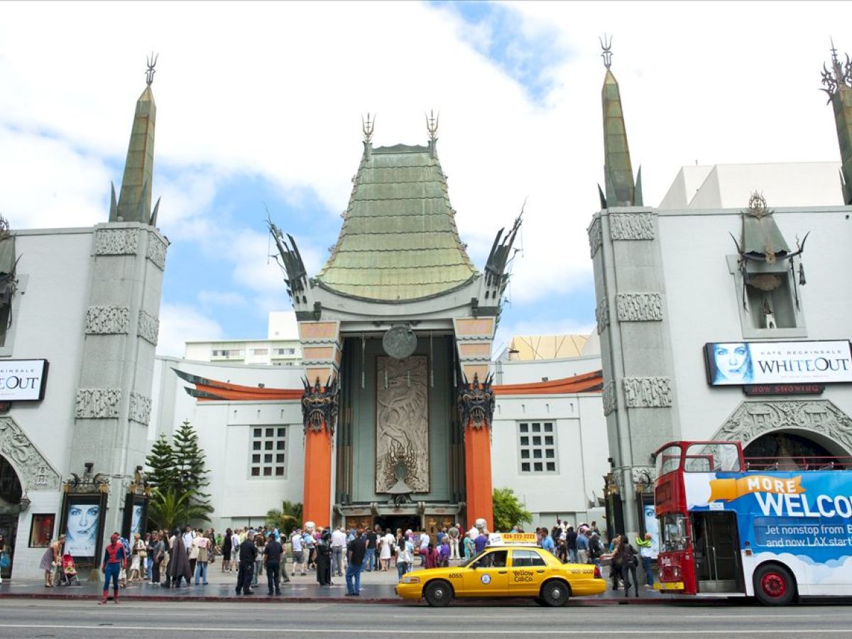 The image shows a famous theater with people, a double-decker bus, and a taxi in front of it, alongside advertisements for a movie named 