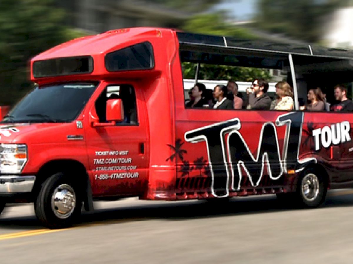 A red TMZ tour bus is shown driving down the street with passengers seated inside, observing attractions and landmarks.