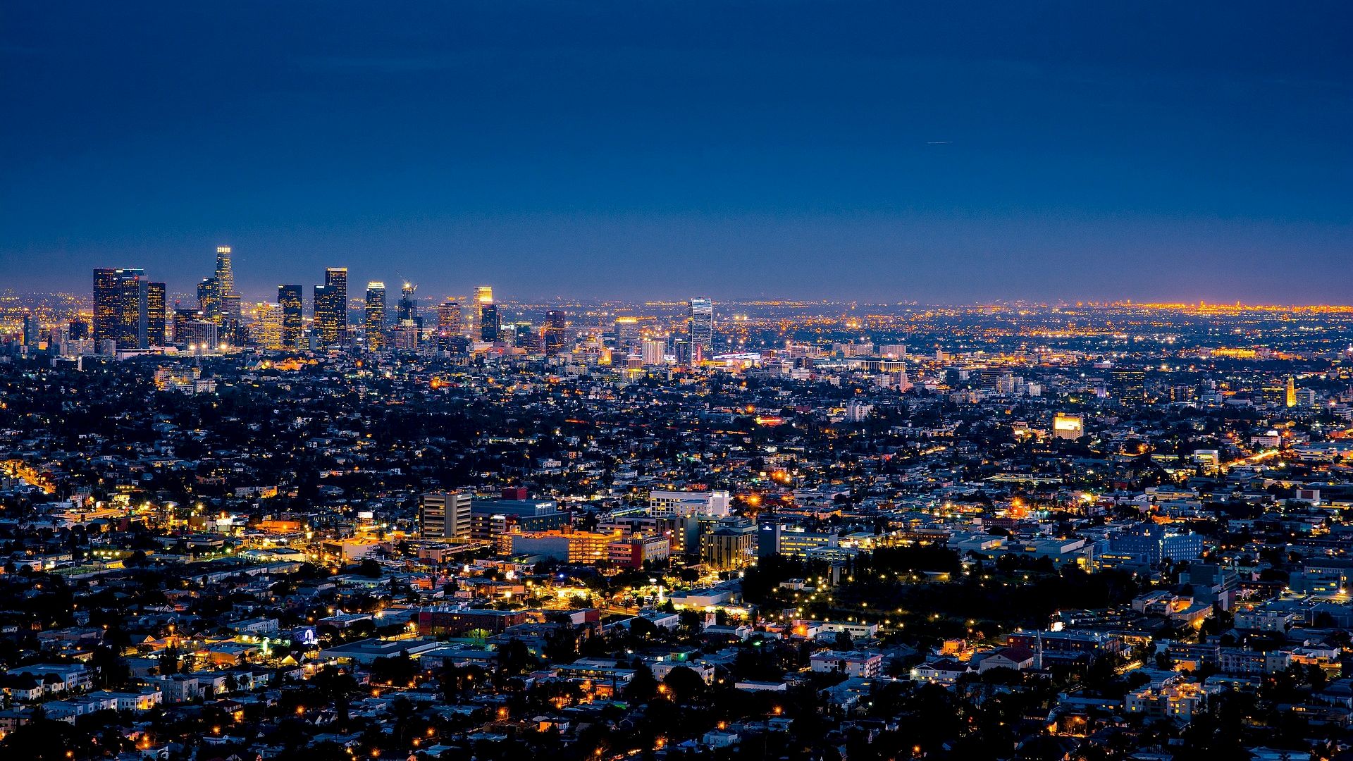 The image shows an aerial night view of a sprawling cityscape with numerous illuminated buildings and lights under a dark blue sky.