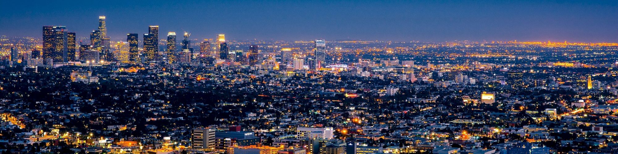 The image shows an aerial night view of a sprawling cityscape with numerous illuminated buildings and lights under a dark blue sky.