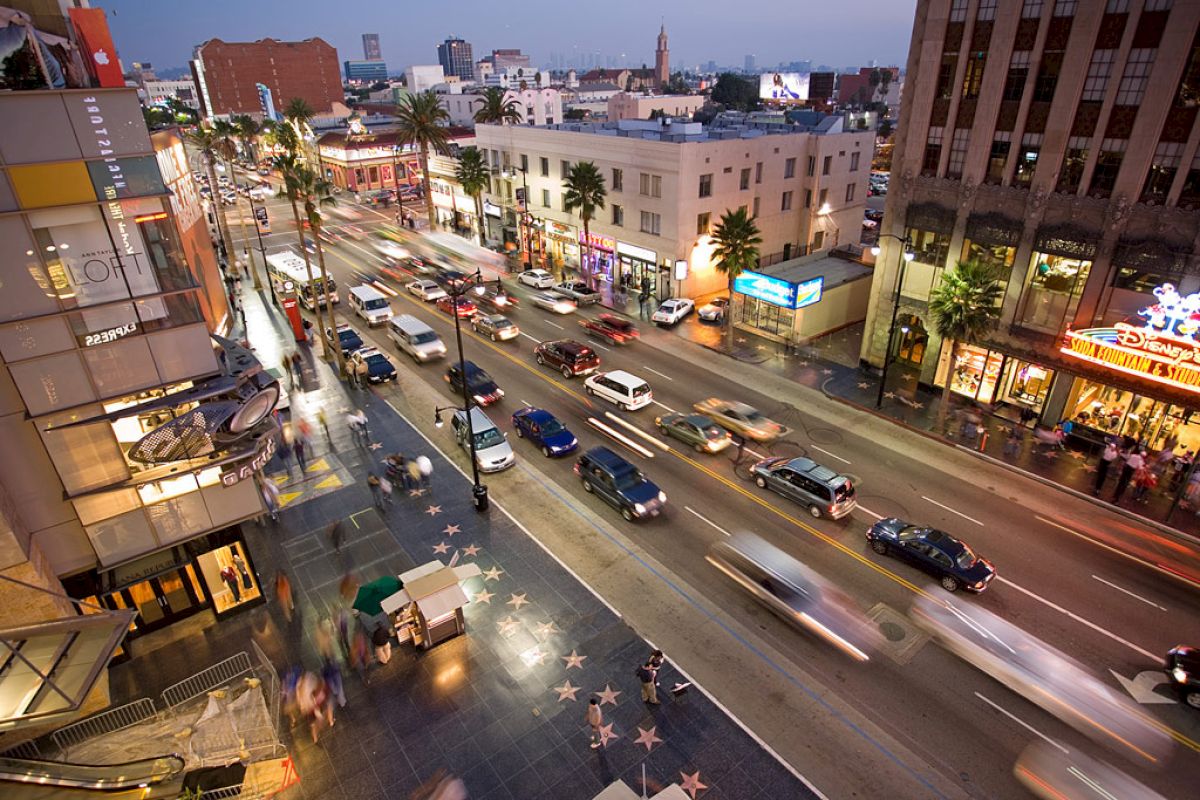 The image shows a busy city street at dusk with moving cars, buildings with neon signs, palm trees, and the Hollywood Walk of Fame visible on the sidewalk.