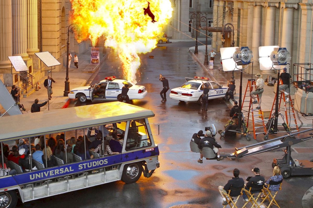 A film set with an explosion scene being filmed, featuring police cars, crew, cameras, and a Universal Studios tour tram with visitors watching.