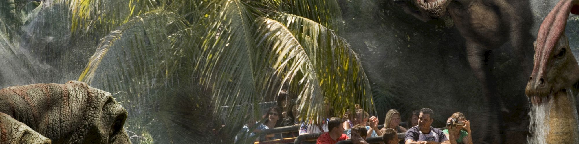 A group of people in a yellow raft navigates a water ride surrounded by large, animatronic dinosaurs in a jungle setting, splashing through water.