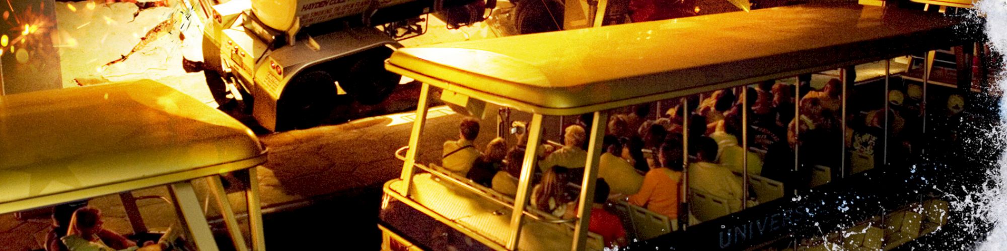 The image shows a theme park ride with people in a tram as pyrotechnic effects and water splashes create a dramatic scene.