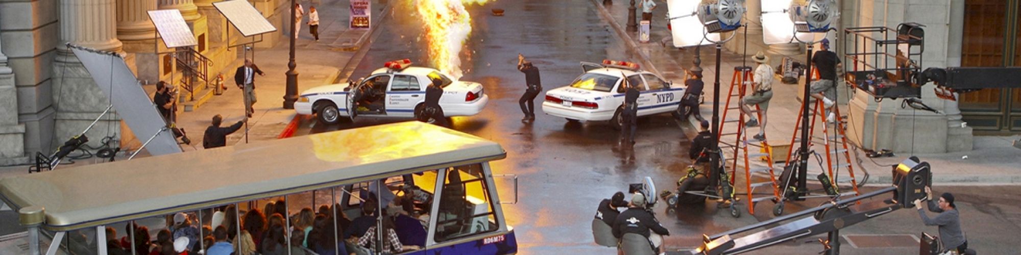 A film set features an explosion, police cars, and crew with lighting equipment as tourists watch from a Universal Studios tram.