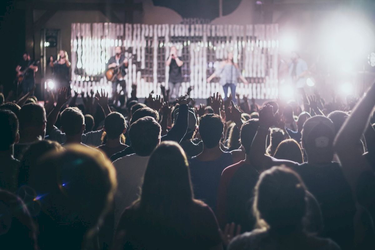 A large crowd is attending a concert with raised hands, while a band performs on stage illuminated by bright lights, creating an energetic atmosphere.