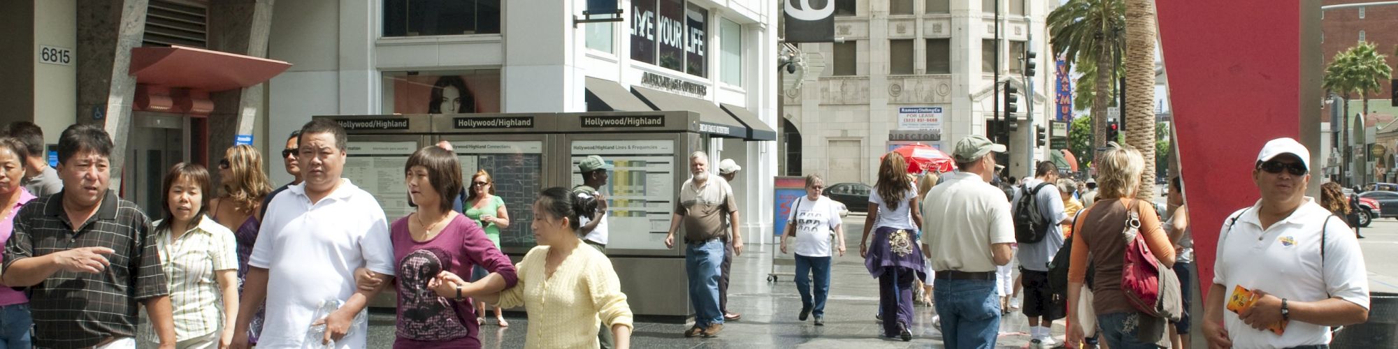 People walking on the Hollywood Walk of Fame, with stars embedded in the sidewalk, and buildings visible in the background, including a Metro station.