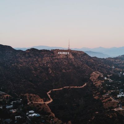 The image shows a landscape view of hilly terrain with the iconic Hollywood Sign in the center, surrounded by sparse buildings and winding roads.