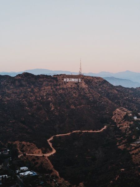 The image shows a landscape view of hilly terrain with the iconic Hollywood Sign in the center, surrounded by sparse buildings and winding roads.