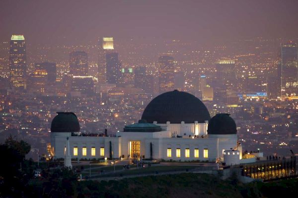 A domed observatory stands on a hill overlooking a cityscape at dusk, with buildings illuminated in the background.