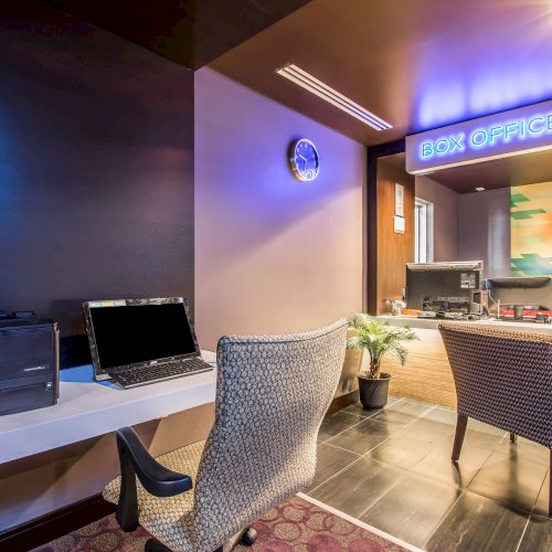 A hotel lobby/check-in area with a &ldquo;Box Office&rdquo; sign, two modern chairs, a desk with a laptop, printer, and a small plant, warm lighting, and a reception counter in the background.