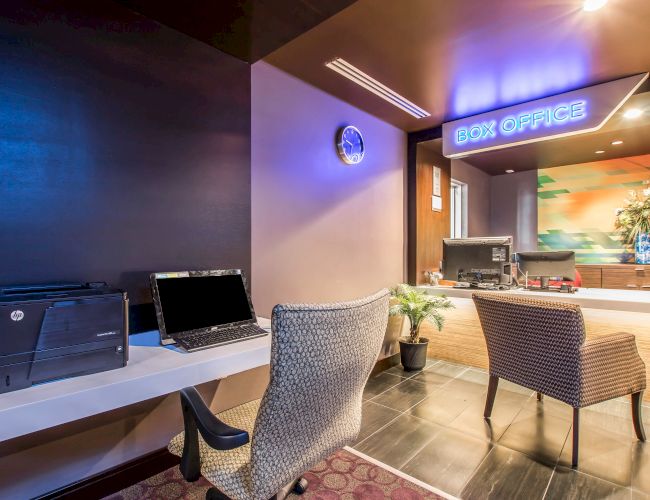 A hotel lobby/check-in area with a &ldquo;Box Office&rdquo; sign, two modern chairs, a desk with a laptop, printer, and a small plant, warm lighting, and a reception counter in the background.