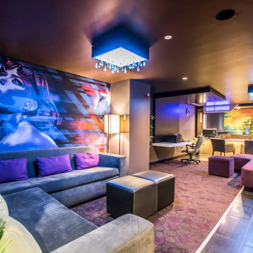 A stylish lounge with a large gray sectional, purple accent pillows, neon-lit bar, colorful wall art featuring a woman, and modern cube ottomans in a cozy, upscale setting.
