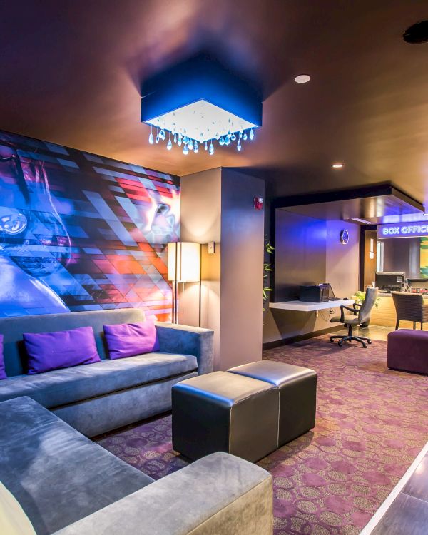 A stylish lounge with a large gray sectional, purple accent pillows, neon-lit bar, colorful wall art featuring a woman, and modern cube ottomans in a cozy, upscale setting.
