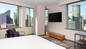 A modern, minimalist hotel room with city skyline views through large windows, featuring a bed, TV, chair, and small furniture.