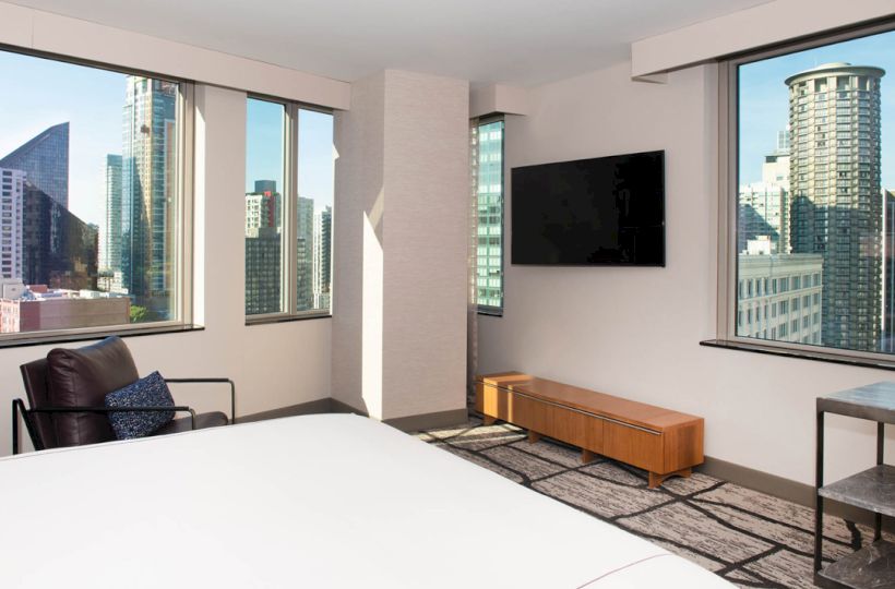 A modern, minimalist hotel room with city skyline views through large windows, featuring a bed, TV, chair, and small furniture.