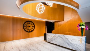 A modern lobby features a large geometric staircase, a marble reception desk, a wall clock, and a vase with flowers.