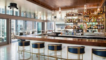 A modern bar with a wooden counter, blue and gold stools, and a wall of various liquor bottles. Large windows offer a cityscape view, ending the sentence.