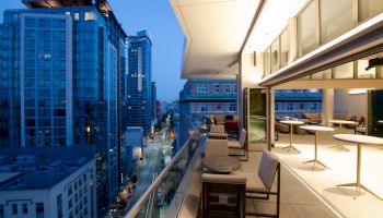 A modern urban balcony at twilight, featuring outdoor seating, glass railings, and views of surrounding skyscrapers and city street below.