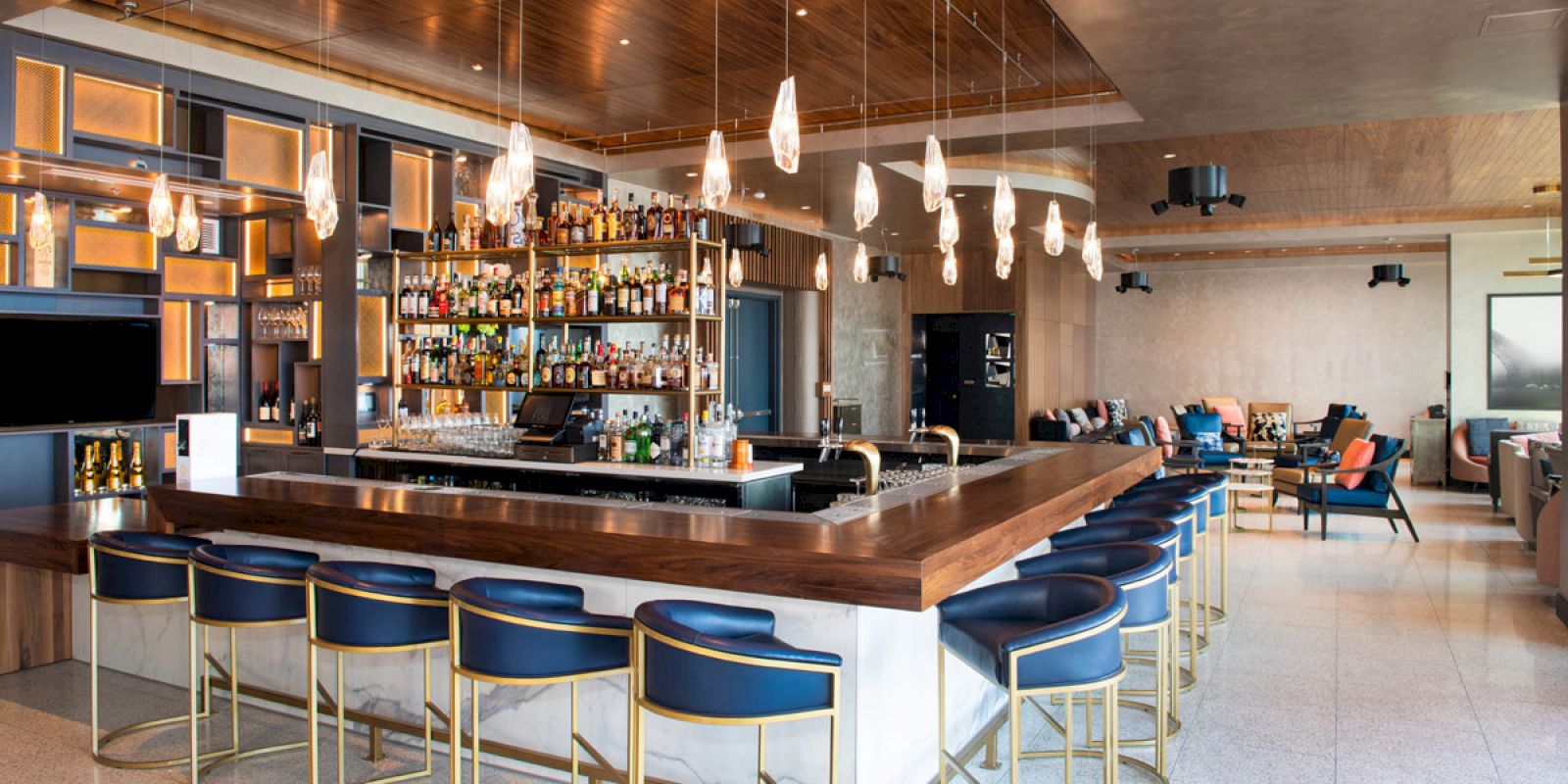 The image shows a modern bar with a central wooden counter, blue bar stools, hanging lights, and shelves stocked with various liquor bottles.