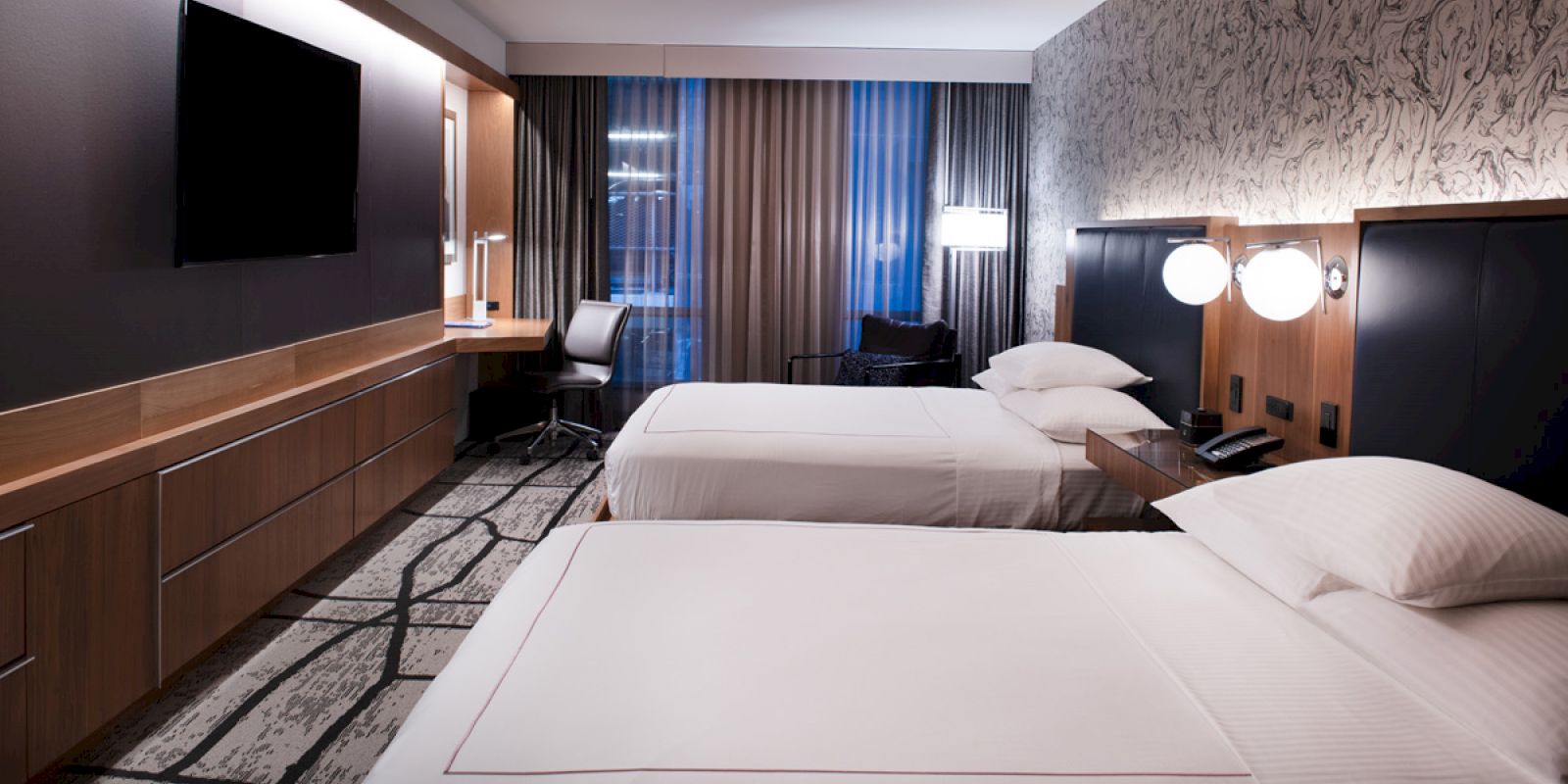 A modern hotel room features two neatly made twin beds, a wall-mounted TV, a desk with a chair, and stylish lighting with patterned wallpaper.