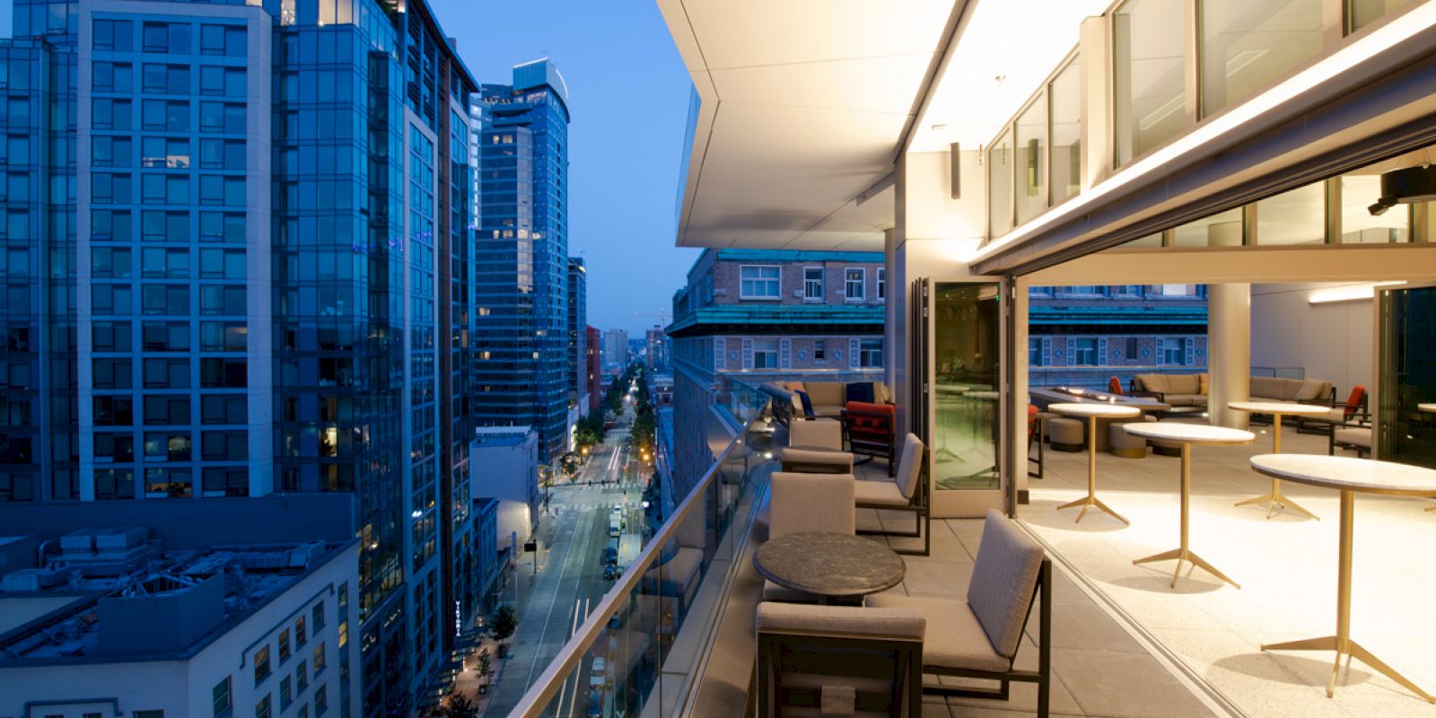 A modern cityscape at dusk, featuring a high-rise balcony with tables and chairs, overlooking illuminated buildings and an urban street.
