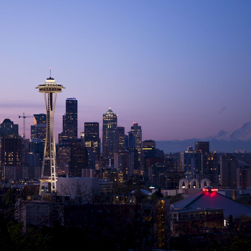 The image shows the Seattle skyline at dusk, highlighting the Space Needle and Mount Rainier in the background, with city lights illuminating the scene.