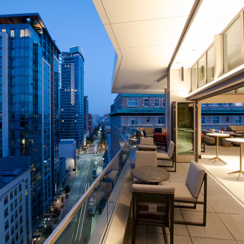 A modern balcony view overlooking a cityscape at dusk, featuring tables and chairs on the balcony and buildings illuminated by city lights.